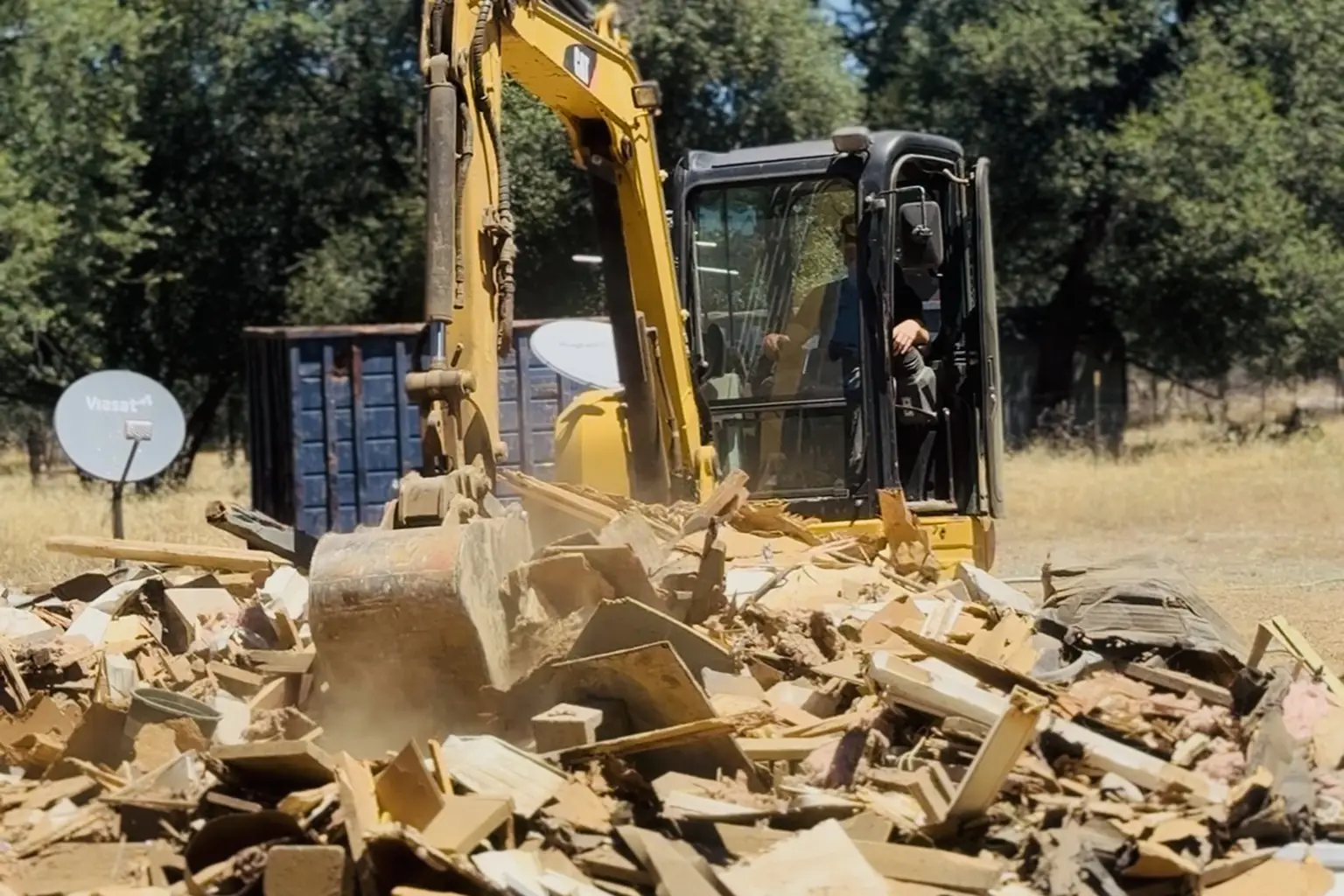 Excavator clearing a mobile home debris pile beside a dump trailer